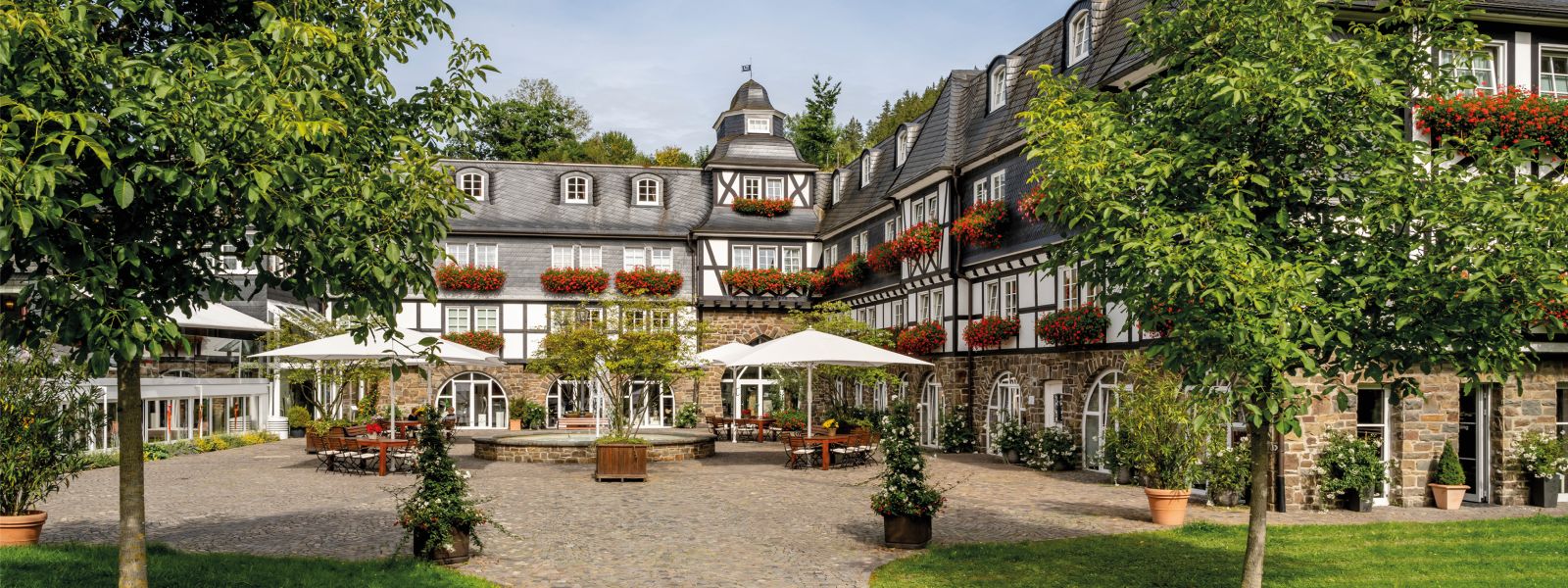 Blick auf einen Fachwerk-Gutshof mit roten Geranien in den Blumenkästen an den Fenstern, im Innenhof stehen Tische, Stühle, Sonnenschirme