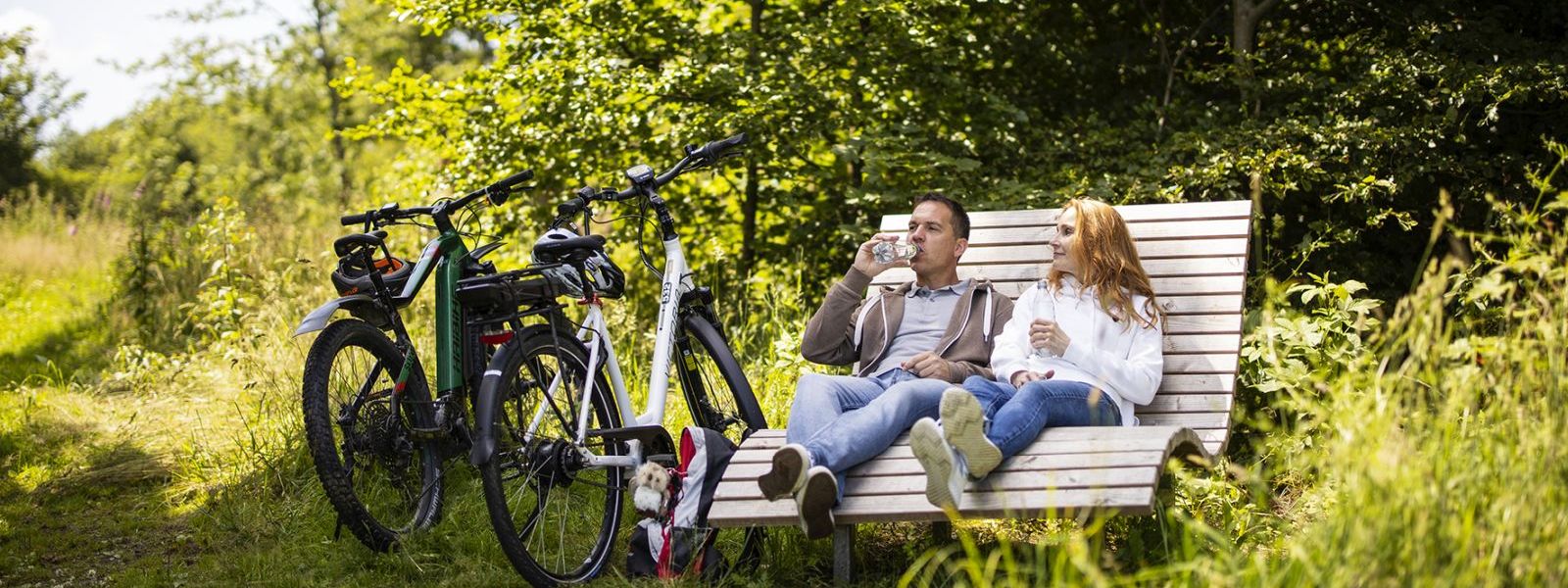 Ein Paar auf Radtour macht auf einer Waldliegebank Pause und trinkt Wasser.