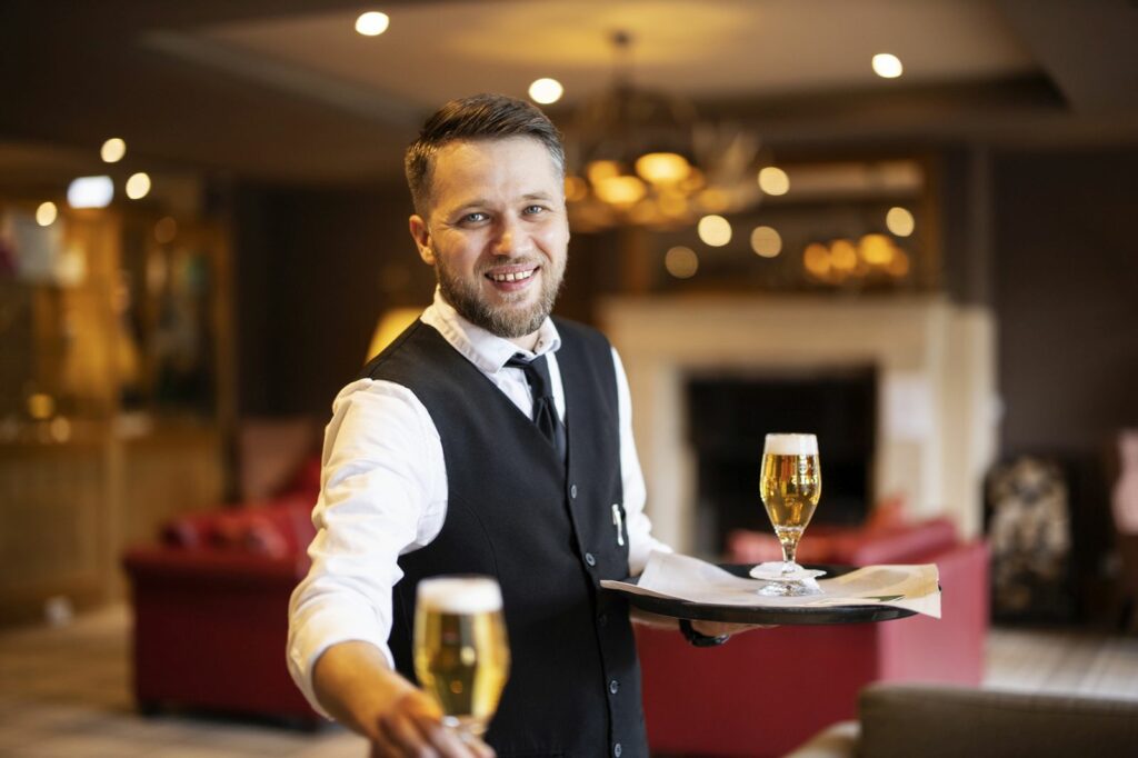 Servicemitarbeiter mit Hemd und Weste; er trägt Tablett mit einem gezapften Bier und reicht ein Glas quasi an den Fotografen