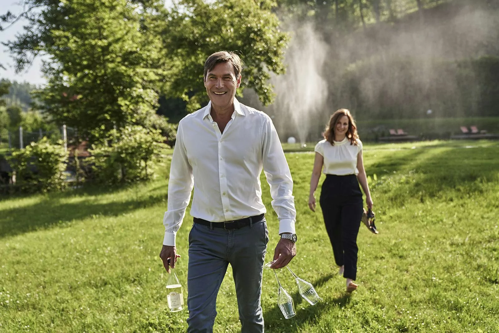 Auf der Suche nach einem Platz im Garten des Hotels Deimann Eine Paar läuft über eine Wiese im Garten vom Hotel Deimann, sie läuft Barfuß, er hält 2 Weingläser und 1 Flasche Wein in den Händen