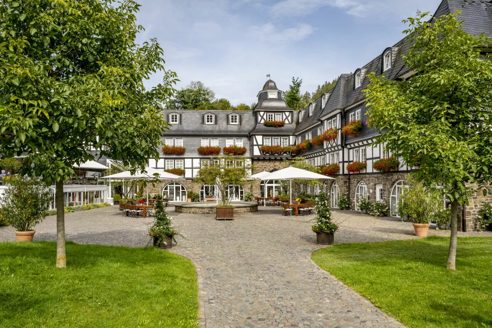 Hotel Deimann – Gutshof Blick auf einen Fachwerk-Gutshof mit roten Geranien in den Blumenkästen an den Fenstern, im Innenhof stehen Tische, Stühle, Sonnenschirme