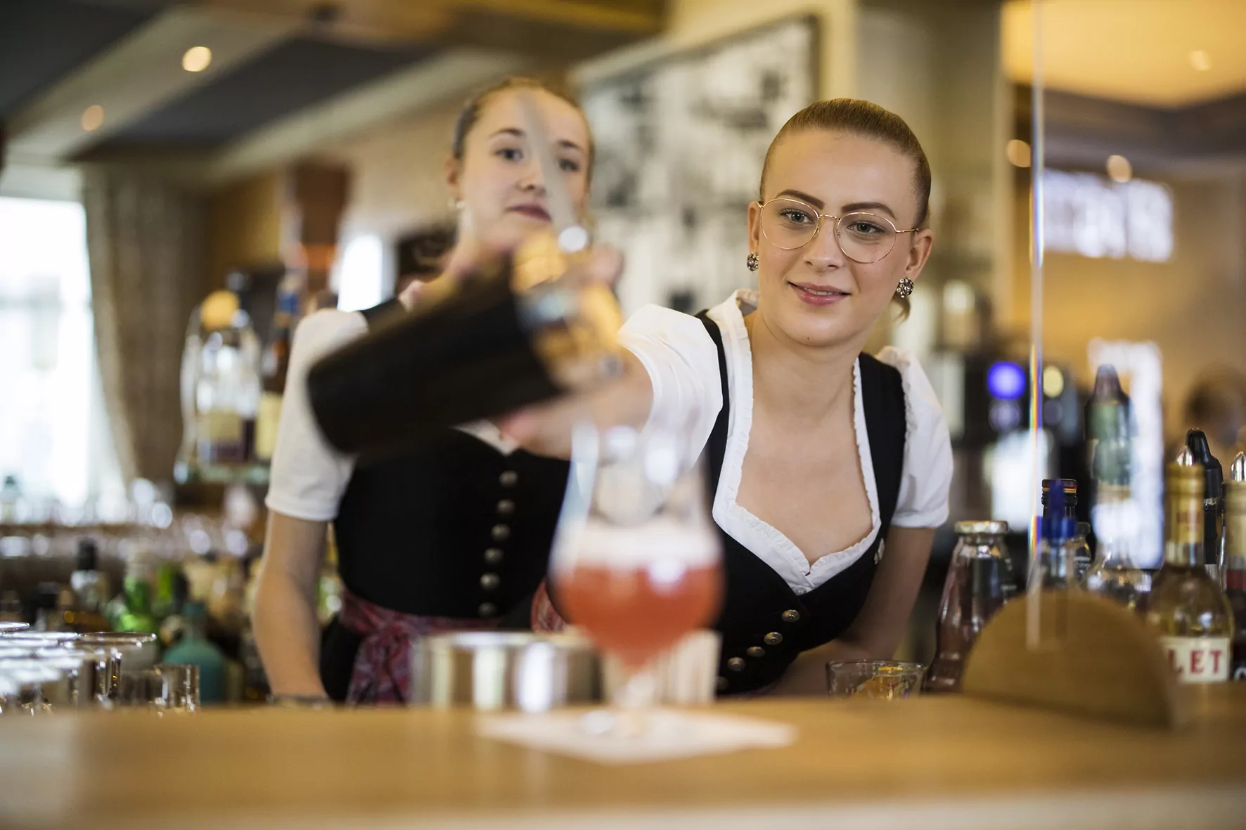 Eine Kollegin in der Bar schenkt einen Cocktail ein; ein Azubi schaut ihr über die Schulter.