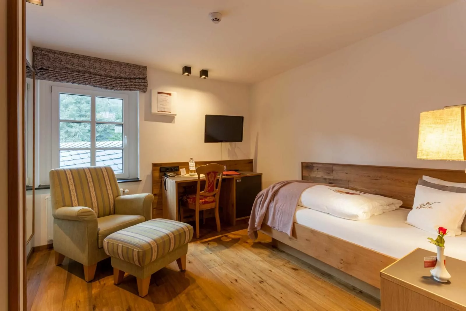 Landhotel Voss – gemütliches Einzelzimmer Einzelzimmer vom Hotel Voss mit Einzelbett in der Ecke und Sessel mit Fußbänkchen sowie Schreibtisch und Nachttisch